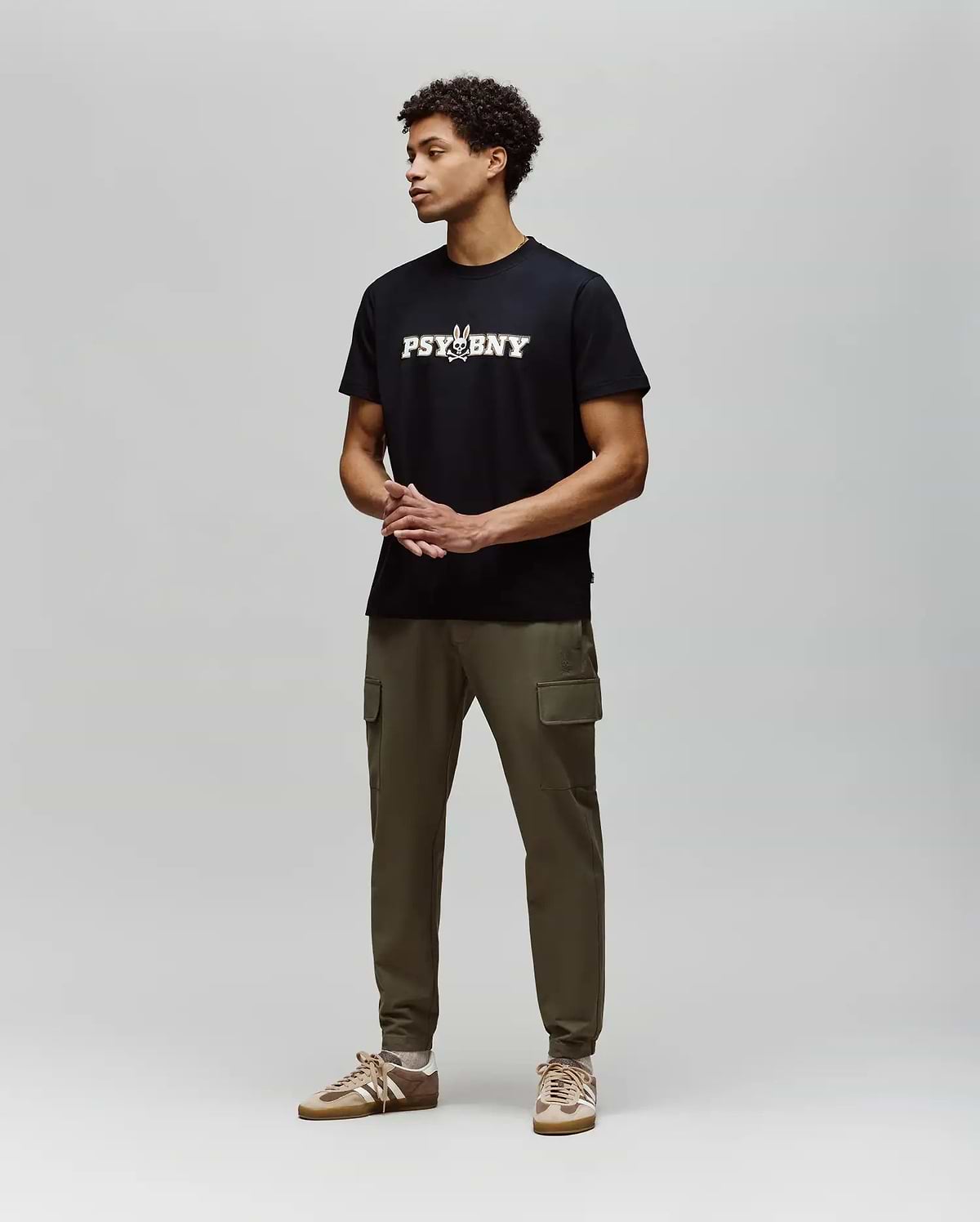 A man stands against a light gray background, wearing the Psycho Bunny MENS WADE GRAPHIC TEE - MT0200105 in black with olive green cargo pants and tan sneakers. His hands are clasped in front as he looks to the side. (4)
