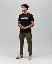 A man stands against a light gray background, wearing the Psycho Bunny MENS WADE GRAPHIC TEE - MT0200105 in black with olive green cargo pants and tan sneakers. His hands are clasped in front as he looks to the side. (4)