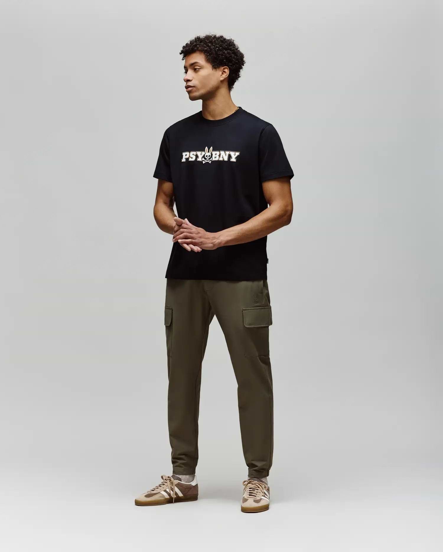 A man stands against a light gray background, wearing the Psycho Bunny MENS WADE GRAPHIC TEE - MT0200105 in black with olive green cargo pants and tan sneakers. His hands are clasped in front as he looks to the side. (4)