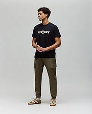 A man stands against a light gray background, wearing the Psycho Bunny MENS WADE GRAPHIC TEE - MT0200105 in black with olive green cargo pants and tan sneakers. His hands are clasped in front as he looks to the side. (4)