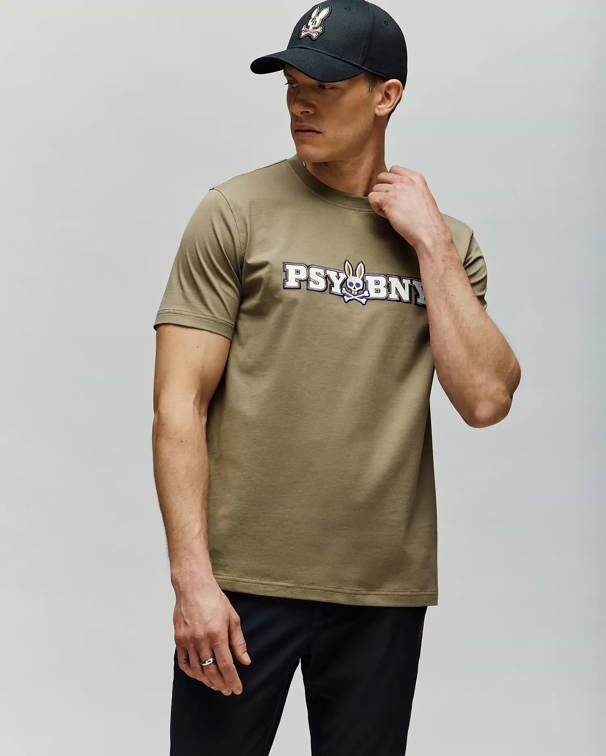 A man in a black cap and the Psycho Bunny MENS WADE GRAPHIC TEE (MT0200105)—olive-green with "PSYBUNY" and a cartoon bunny graphic—stands against a light gray background, looking sideways with one hand on his neck. (4)
