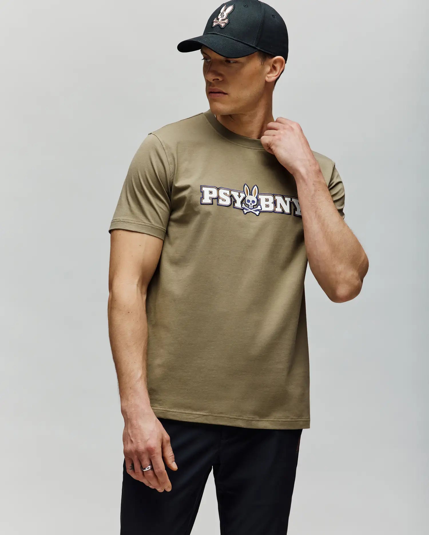 A man in a black cap and the Psycho Bunny MENS WADE GRAPHIC TEE (MT0200105)—olive-green with "PSYBUNY" and a cartoon bunny graphic—stands against a light gray background, looking sideways with one hand on his neck. (4)