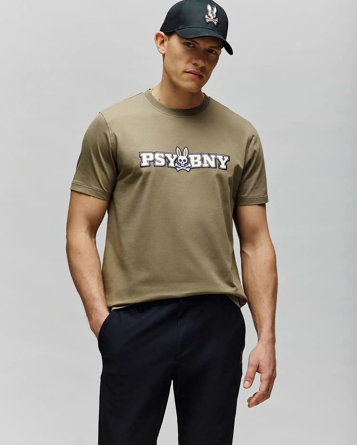 A man in a regular fit, 100% cotton olive green MENS WADE GRAPHIC TEE - MT0200105 by Psycho Bunny with a "PSYBNY" and cartoon bunny logo stands with one hand in his pocket, wearing a black cap against a light gray background. (2)