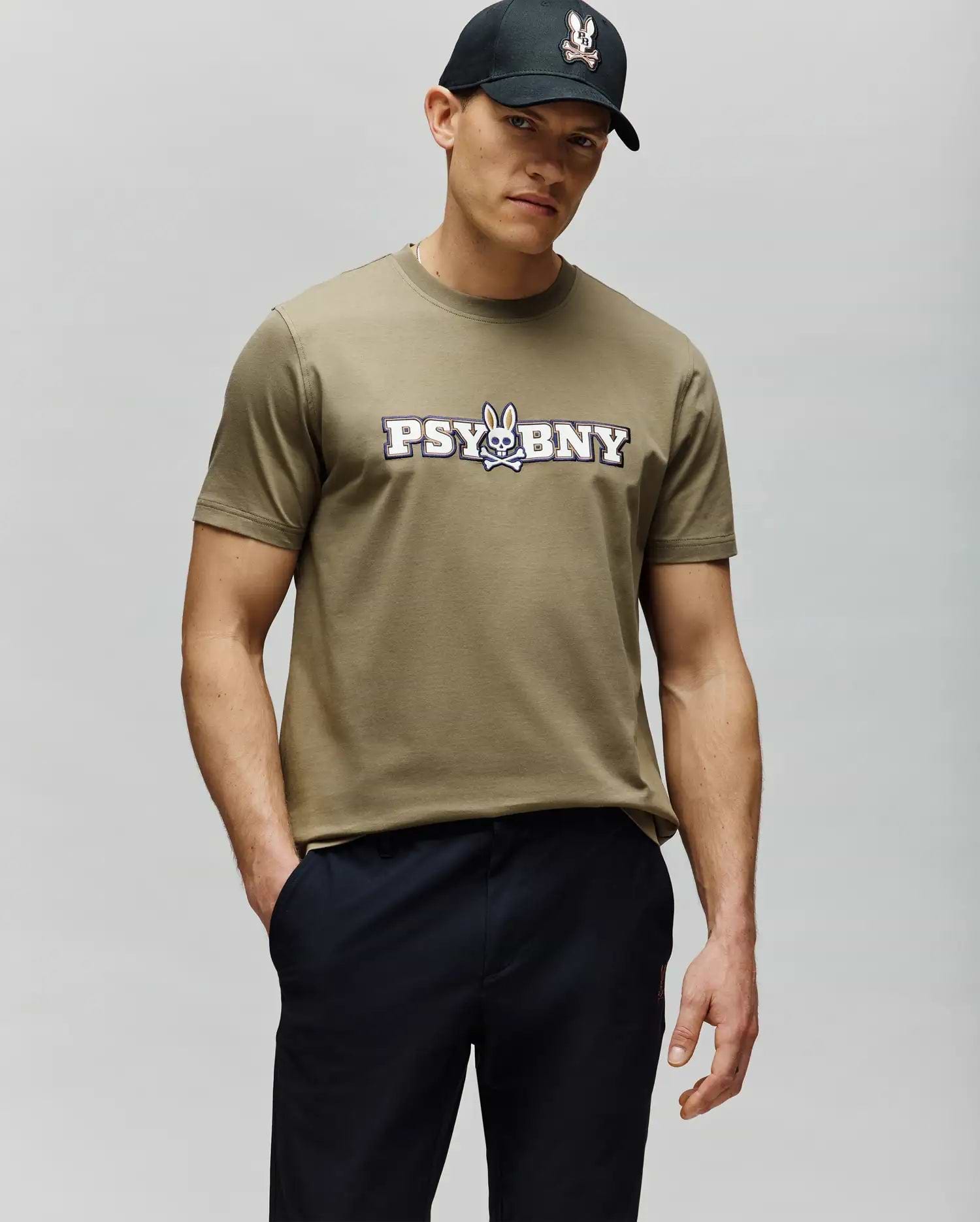 A man in a regular fit, 100% cotton olive green MENS WADE GRAPHIC TEE - MT0200105 by Psycho Bunny with a "PSYBNY" and cartoon bunny logo stands with one hand in his pocket, wearing a black cap against a light gray background. (2)