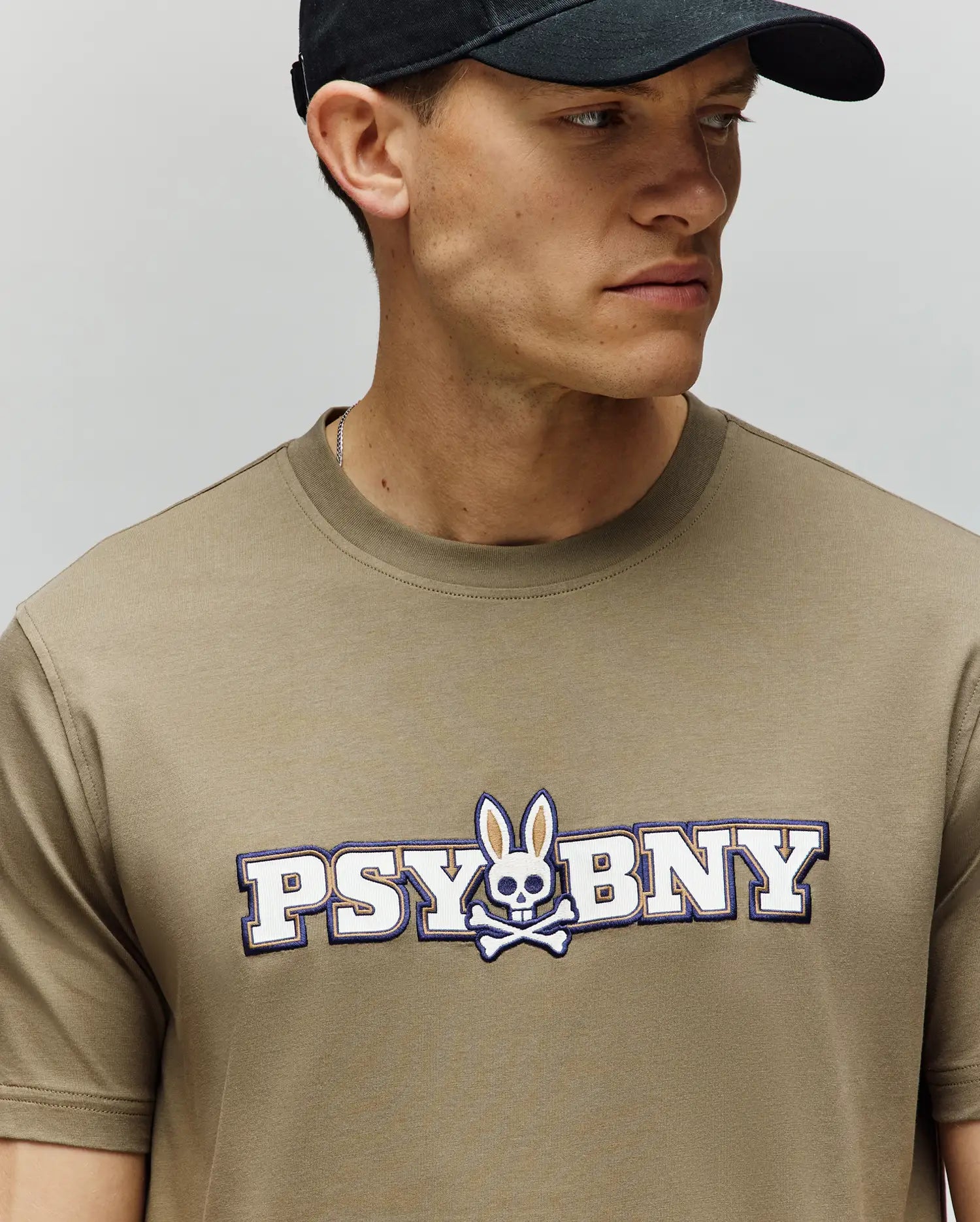 A man wears a Psycho Bunny MENS WADE GRAPHIC TEE (MT0200105) in beige, featuring "PSYBNY" and a bunny skull and crossbones on the chest, made from 100% cotton in a regular fit, paired with a black cap, looking to his left. (3)