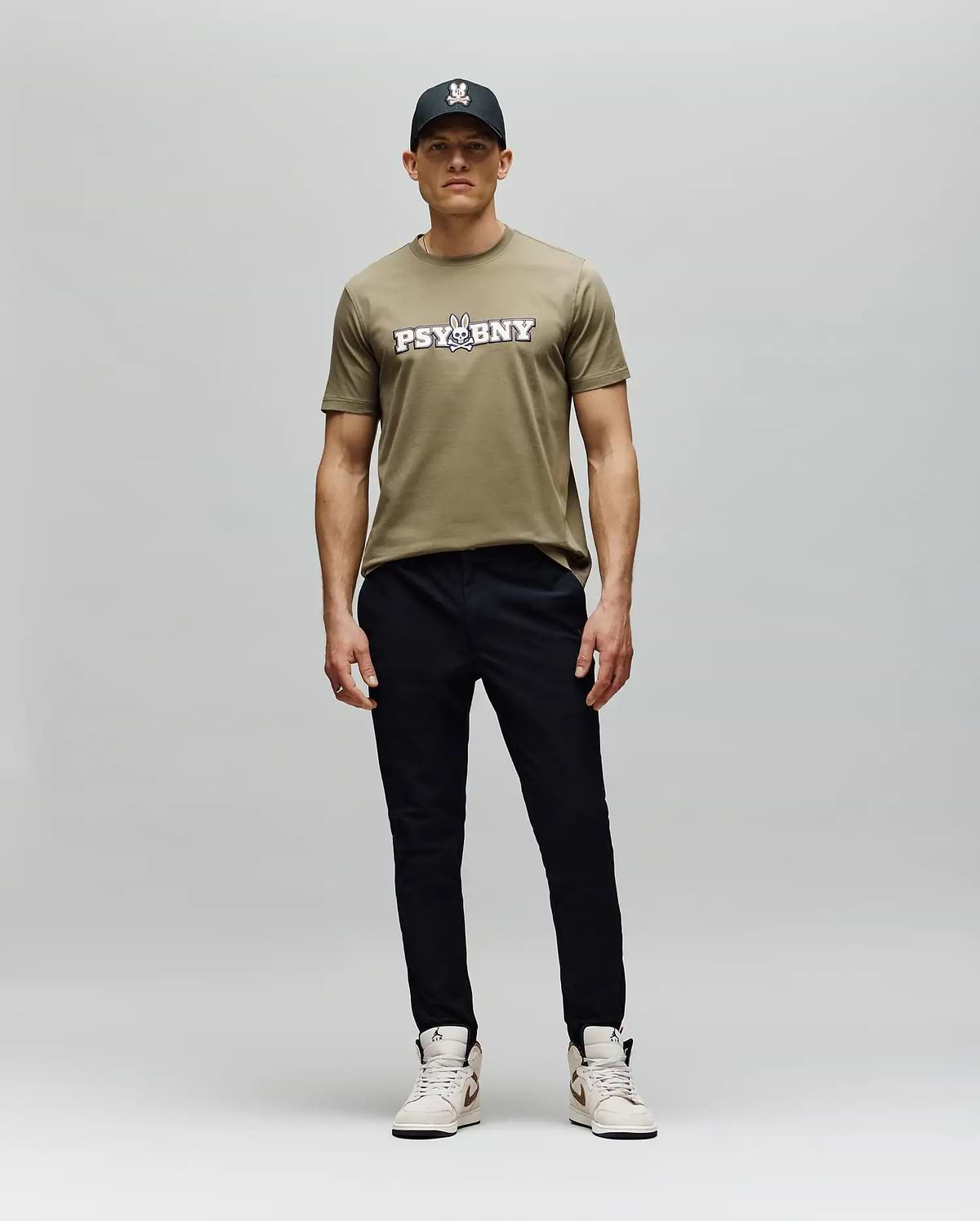 A man stands against a plain background in the Psycho Bunny MENS WADE GRAPHIC TEE - MT0200105, a khaki 100% cotton regular fit t-shirt, paired with a black cap, black pants, and light sneakers. He faces forward with a neutral expression. (5)