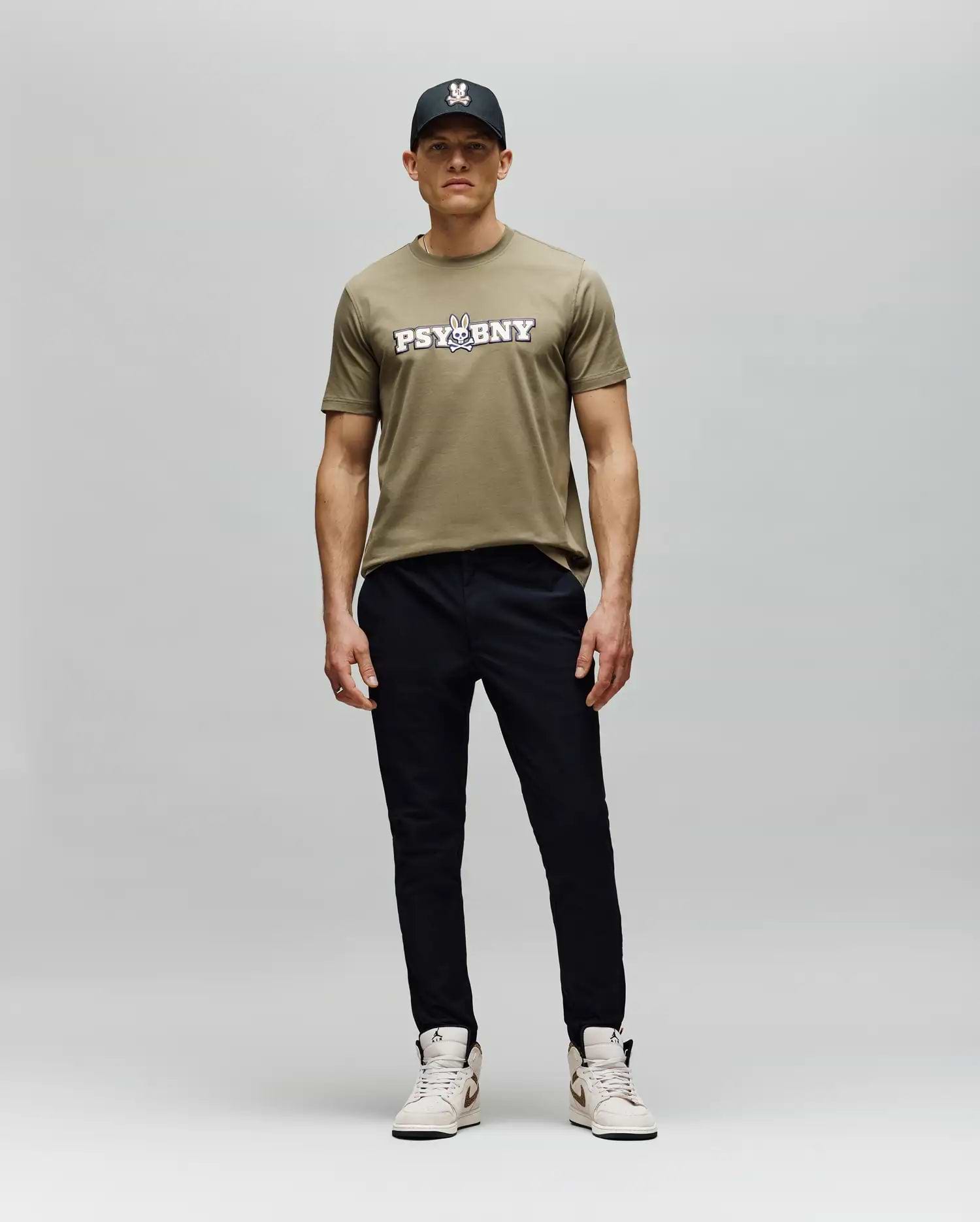 A man stands against a plain background in the Psycho Bunny MENS WADE GRAPHIC TEE - MT0200105, a khaki 100% cotton regular fit t-shirt, paired with a black cap, black pants, and light sneakers. He faces forward with a neutral expression. (5)
