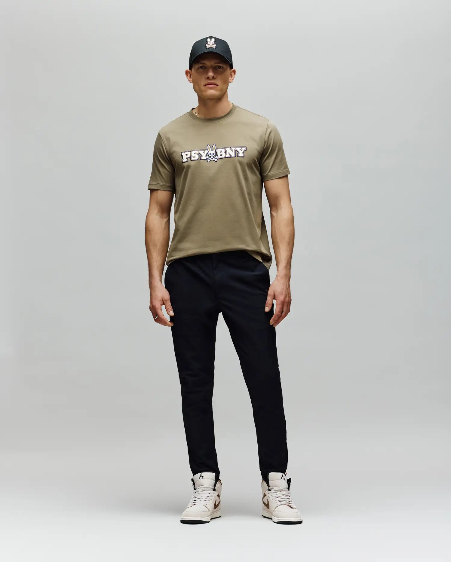 A man stands against a plain background in the Psycho Bunny MENS WADE GRAPHIC TEE - MT0200105, a khaki 100% cotton regular fit t-shirt, paired with a black cap, black pants, and light sneakers. He faces forward with a neutral expression. (5)