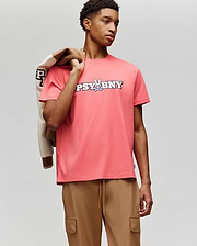A young man wears the Psycho Bunny MENS WADE GRAPHIC TEE - MT0200105, a regular fit 100% cotton pink tee featuring "PSYBNY" and a cartoon bunny, paired with tan cargo pants and holding a beige and brown jacket. (2)