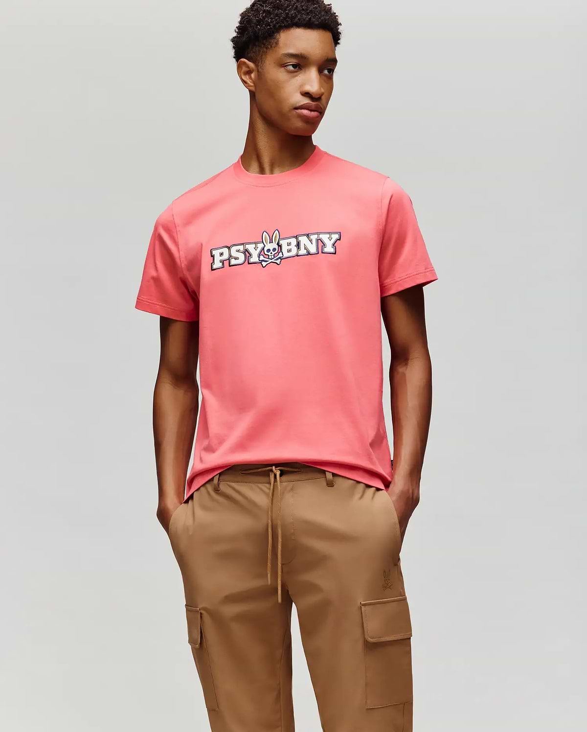 A person wears the Psycho Bunny MENS WADE GRAPHIC TEE - MT0200105, a coral pink 100% cotton tee with "PSYBNY" and a cartoon bunny on the chest, paired with tan cargo pants against a light gray background. (4)