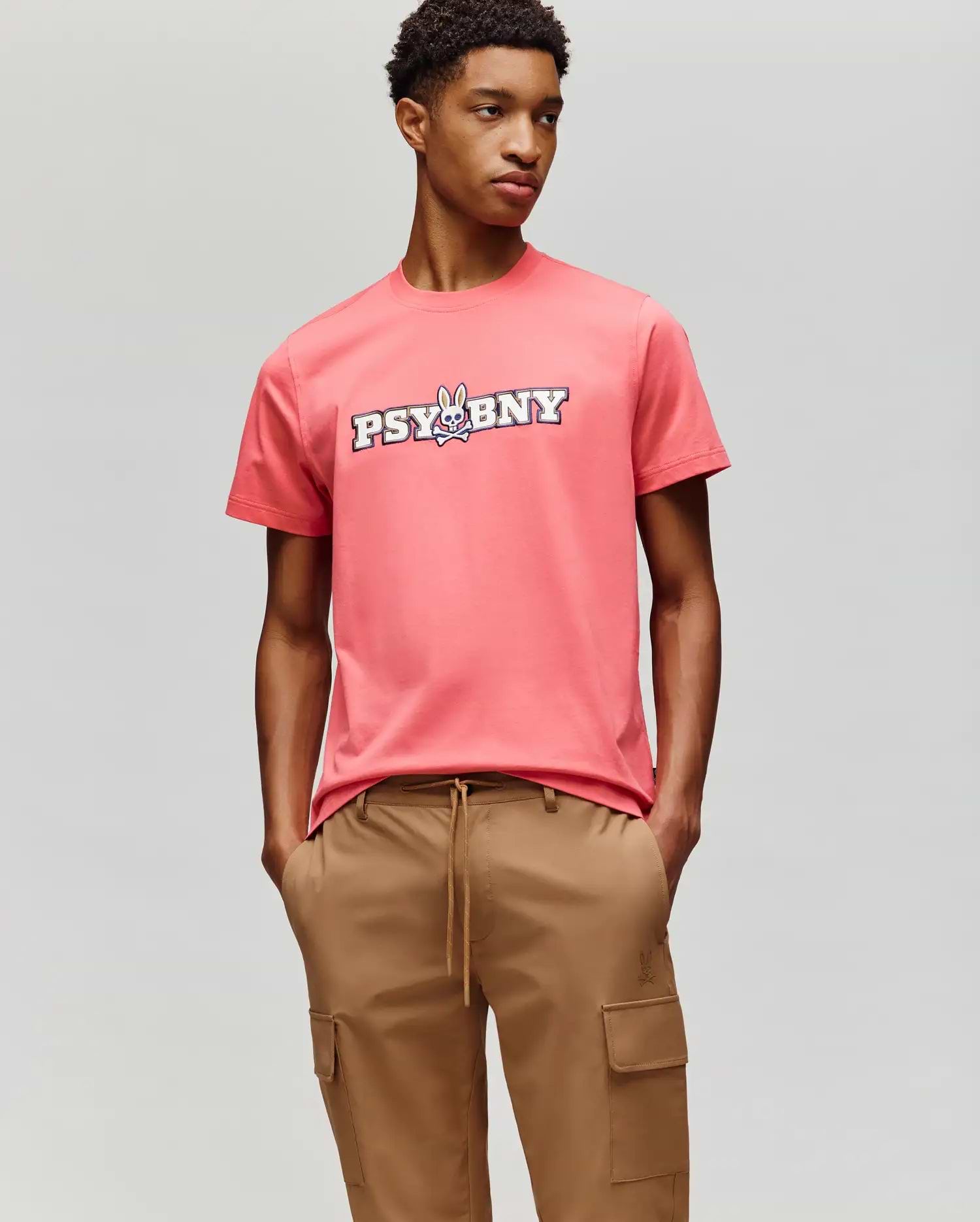 A person wears the Psycho Bunny MENS WADE GRAPHIC TEE - MT0200105, a coral pink 100% cotton tee with "PSYBNY" and a cartoon bunny on the chest, paired with tan cargo pants against a light gray background. (4)