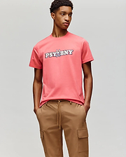 A person wears the Psycho Bunny MENS WADE GRAPHIC TEE - MT0200105, a coral pink 100% cotton tee with "PSYBNY" and a cartoon bunny on the chest, paired with tan cargo pants against a light gray background. (4)