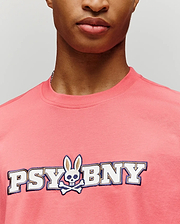 A person in the Psycho Bunny MENS WADE GRAPHIC TEE (MT0200105), a pink 100% cotton regular fit, featuring "PSYBNY" text and a bunny with skull face and crossbones, shown from shoulders to just below the nose. (3)