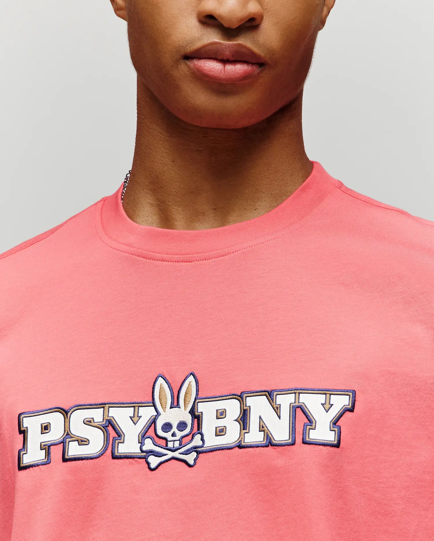 A person in the Psycho Bunny MENS WADE GRAPHIC TEE (MT0200105), a pink 100% cotton regular fit, featuring "PSYBNY" text and a bunny with skull face and crossbones, shown from shoulders to just below the nose. (3)