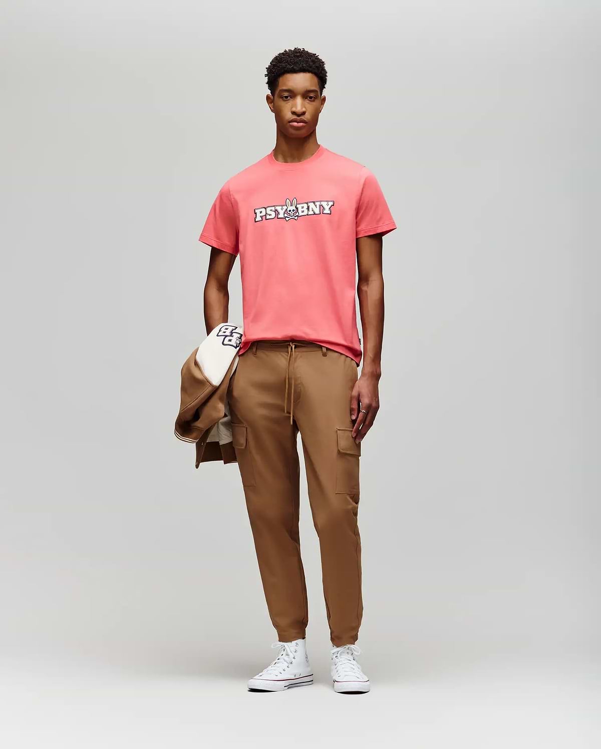 A young man faces the camera in a coral Psycho Bunny MENS WADE GRAPHIC TEE (MT0200105), tan cargo joggers, and white sneakers, holding a tan jacket and cap against a plain light gray background. (5)