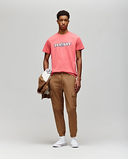 A young man faces the camera in a coral Psycho Bunny MENS WADE GRAPHIC TEE (MT0200105), tan cargo joggers, and white sneakers, holding a tan jacket and cap against a plain light gray background. (5)