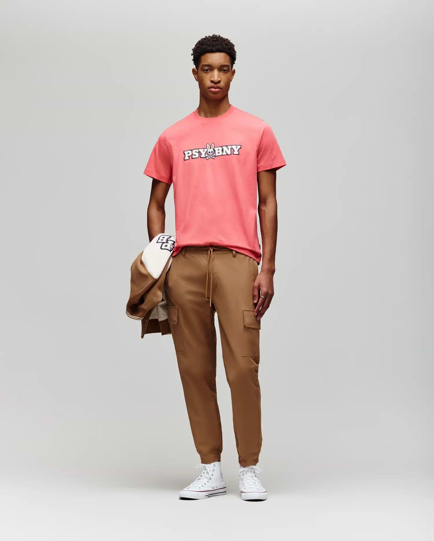 A young man faces the camera in a coral Psycho Bunny MENS WADE GRAPHIC TEE (MT0200105), tan cargo joggers, and white sneakers, holding a tan jacket and cap against a plain light gray background. (5)