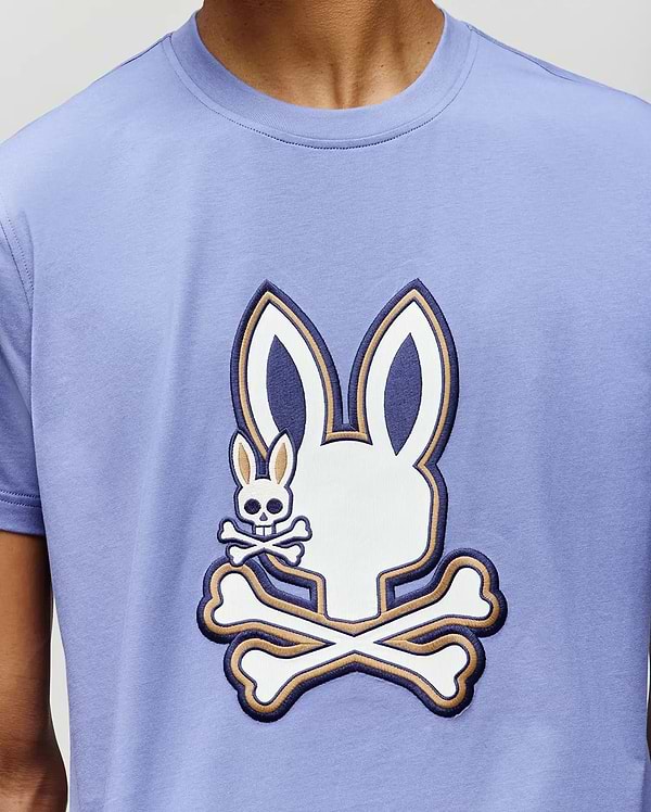 A person wearing the Psycho Bunny MENS WADE GRAPHIC TEE (MT0200106), a light purple 100% cotton shirt with a large white bunny skull and crossbones graphic, plus a smaller bunny skull and crossbones on the cheek.