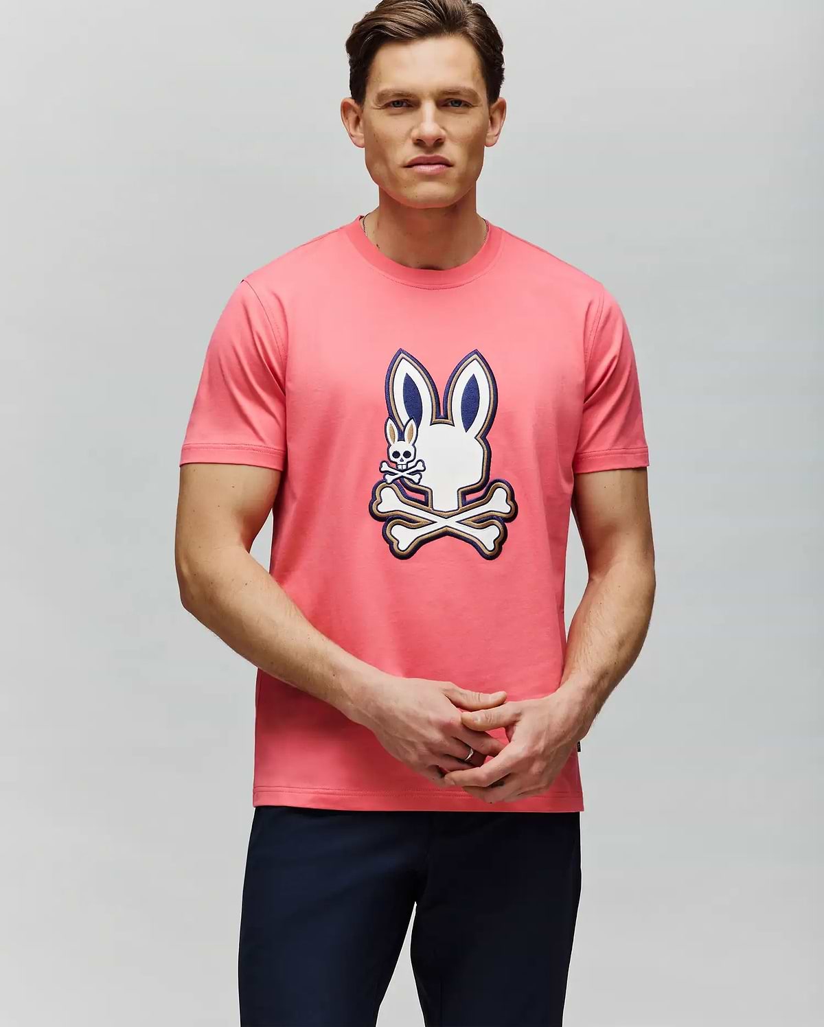 A man with short brown hair and dark pants stands against a light gray backdrop, wearing the Psycho Bunny MENS WADE GRAPHIC TEE - MT0200106, a 100% cotton pink tee featuring an embroidered bunny and crossed bones. (4)