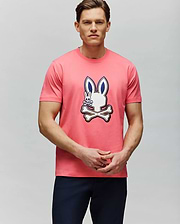 A man with short brown hair and dark pants stands against a light gray backdrop, wearing the Psycho Bunny MENS WADE GRAPHIC TEE - MT0200106, a 100% cotton pink tee featuring an embroidered bunny and crossed bones. (4)