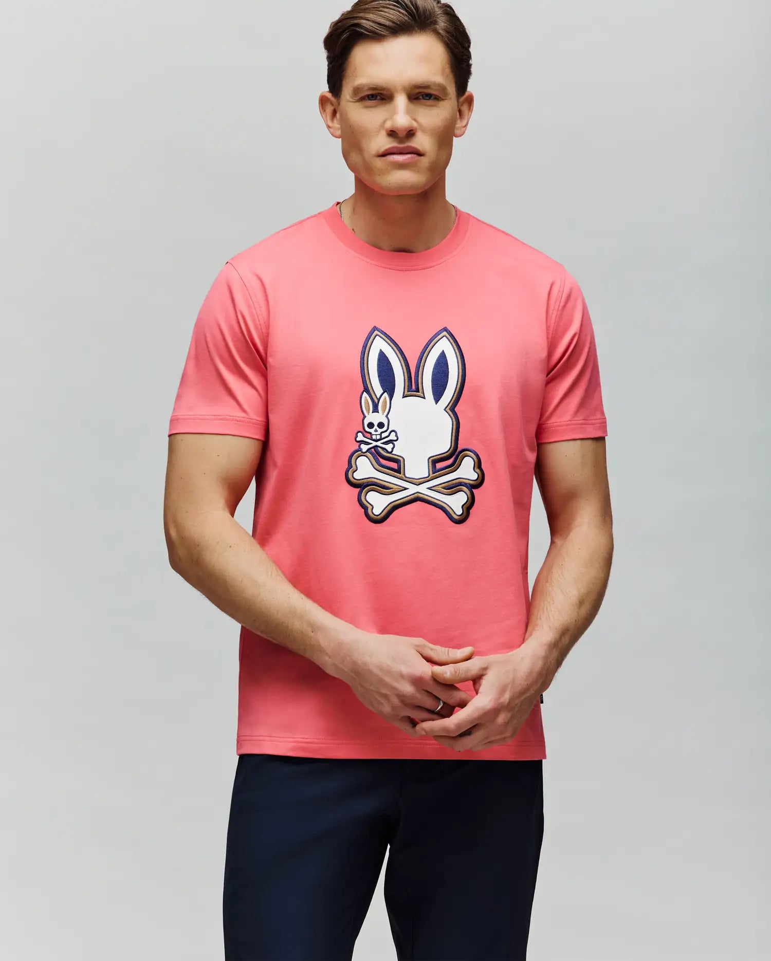 A man with short brown hair and dark pants stands against a light gray backdrop, wearing the Psycho Bunny MENS WADE GRAPHIC TEE - MT0200106, a 100% cotton pink tee featuring an embroidered bunny and crossed bones. (4)