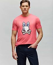 A man wears the Psycho Bunny MENS WADE GRAPHIC TEE - MT0200106, a pink 100% cotton shirt with a cartoon bunny and crossbones graphic on the front, paired with dark pants against a plain light gray background. (2)