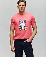 A man wears the Psycho Bunny MENS WADE GRAPHIC TEE - MT0200106, a pink 100% cotton shirt with a cartoon bunny and crossbones graphic on the front, paired with dark pants against a plain light gray background. (2)