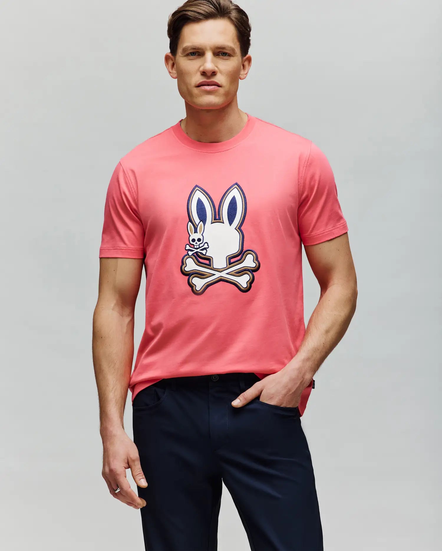A man wears the Psycho Bunny MENS WADE GRAPHIC TEE - MT0200106, a pink 100% cotton shirt with a cartoon bunny and crossbones graphic on the front, paired with dark pants against a plain light gray background. (2)
