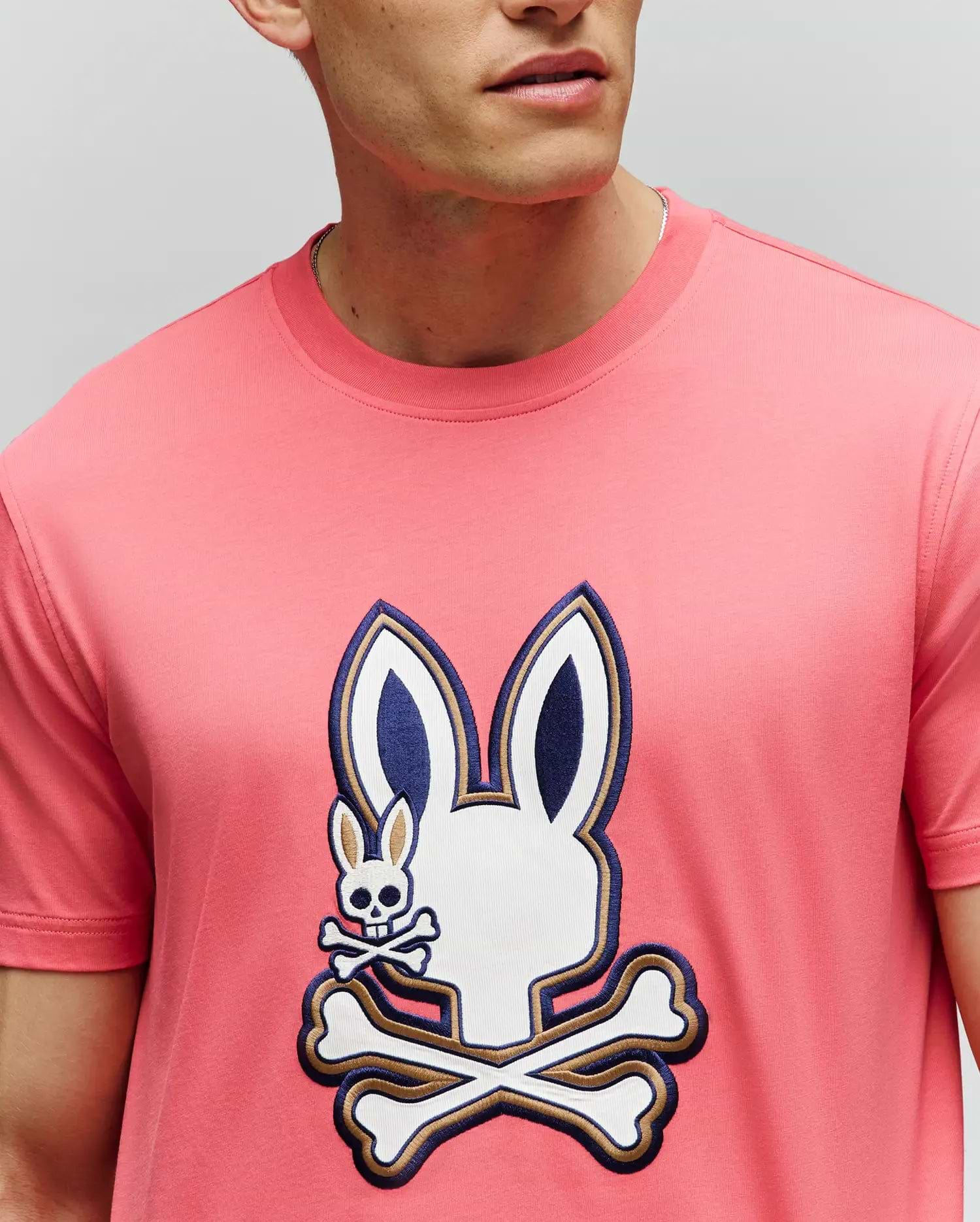 A man wears the Psycho Bunny MENS WADE GRAPHIC TEE (MT0200106), made from 100% cotton, featuring a large bunny head with crossbones graphic and a small embroidered bunny with crossbones on the chest. His face is partially out of frame. (3)