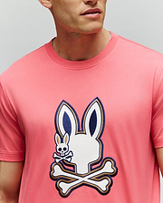A man wears the Psycho Bunny MENS WADE GRAPHIC TEE (MT0200106), made from 100% cotton, featuring a large bunny head with crossbones graphic and a small embroidered bunny with crossbones on the chest. His face is partially out of frame. (3)