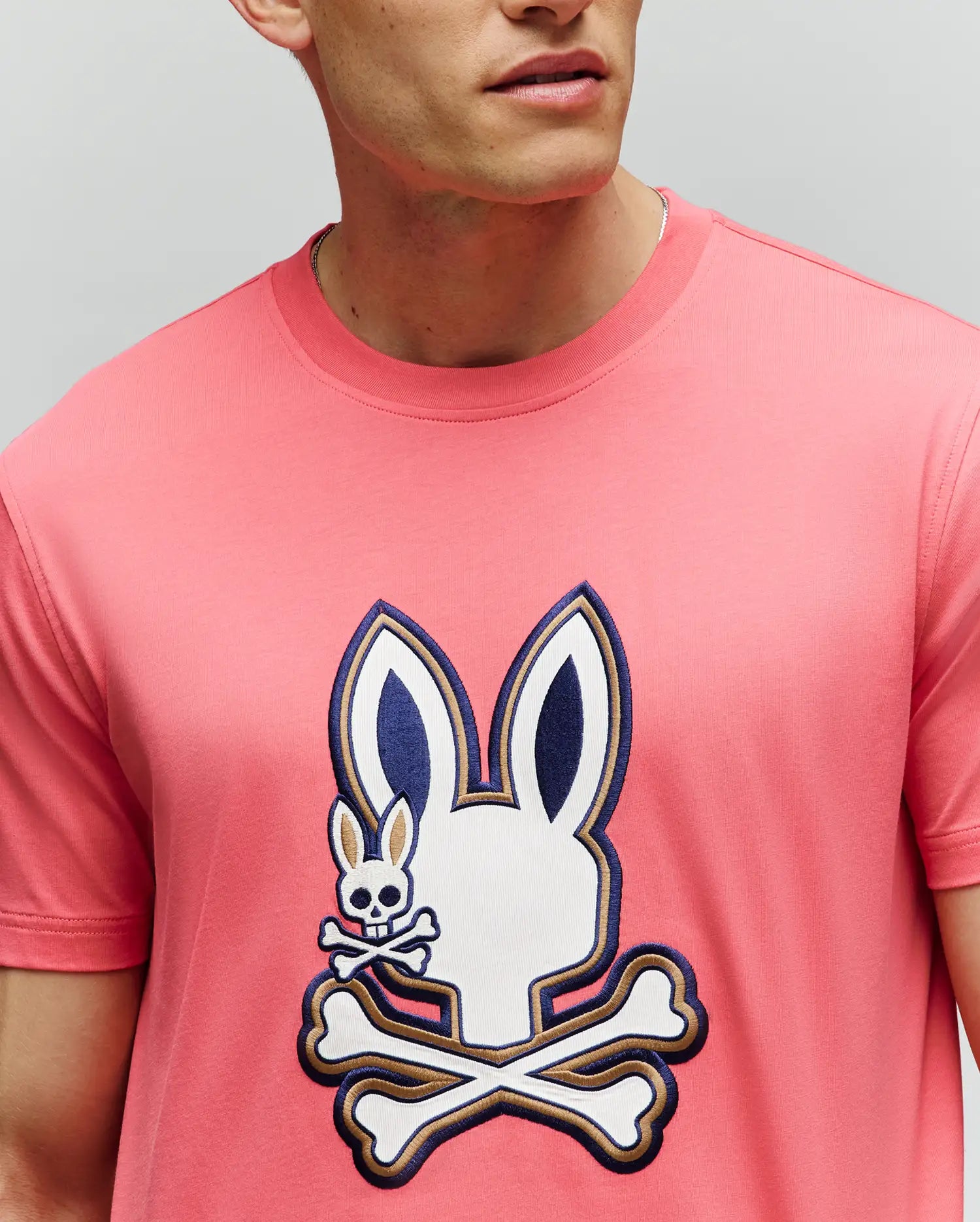 A man wears the Psycho Bunny MENS WADE GRAPHIC TEE (MT0200106), made from 100% cotton, featuring a large bunny head with crossbones graphic and a small embroidered bunny with crossbones on the chest. His face is partially out of frame. (3)
