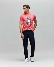 A man poses against a plain background in the Psycho Bunny MENS WADE GRAPHIC TEE (MT0200106), a pink 100% cotton shirt with an embroidered bunny and crossbones, styled with dark pants and white sneakers, one hand in his pocket. (5)