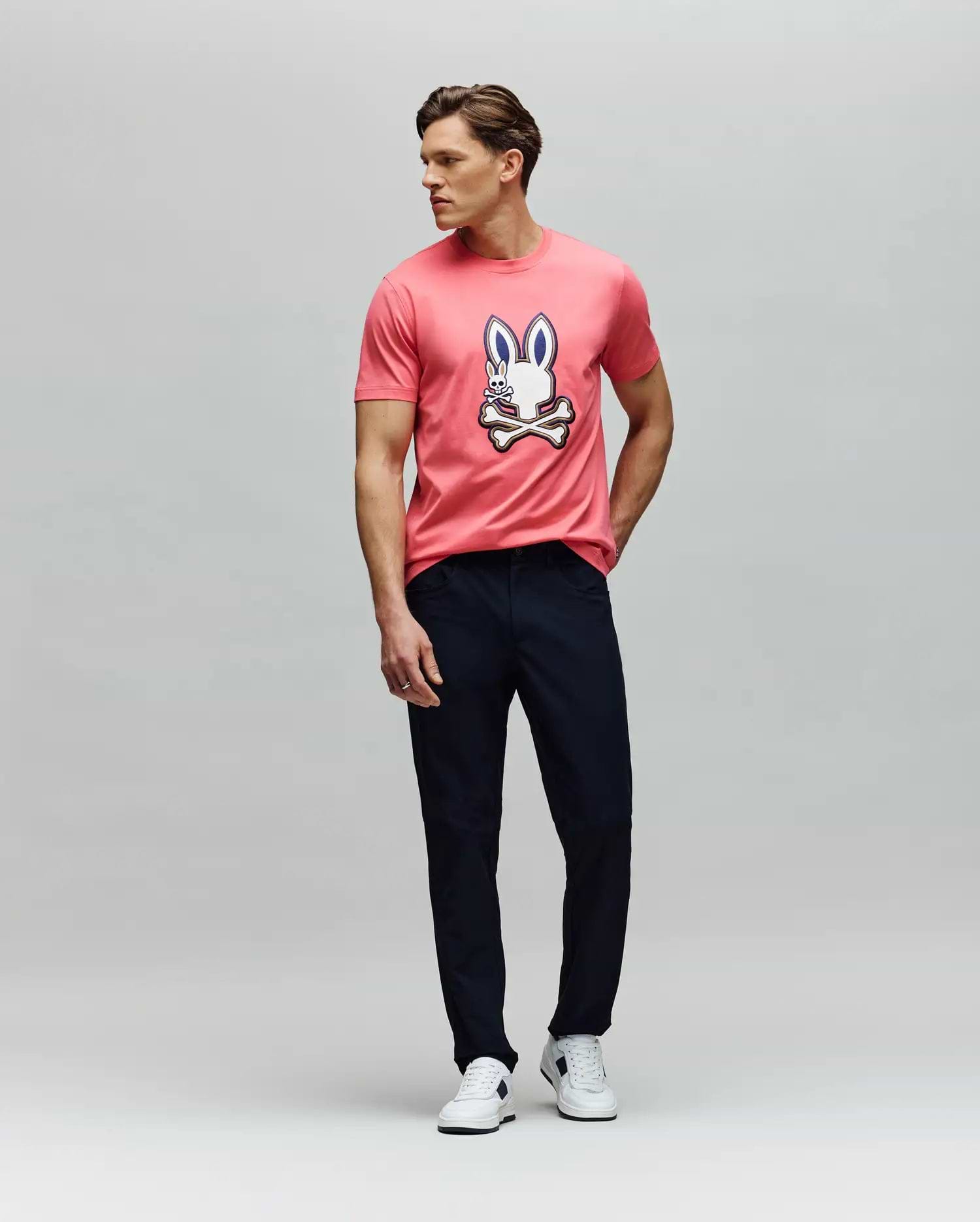 A man poses against a plain background in the Psycho Bunny MENS WADE GRAPHIC TEE (MT0200106), a pink 100% cotton shirt with an embroidered bunny and crossbones, styled with dark pants and white sneakers, one hand in his pocket. (5)