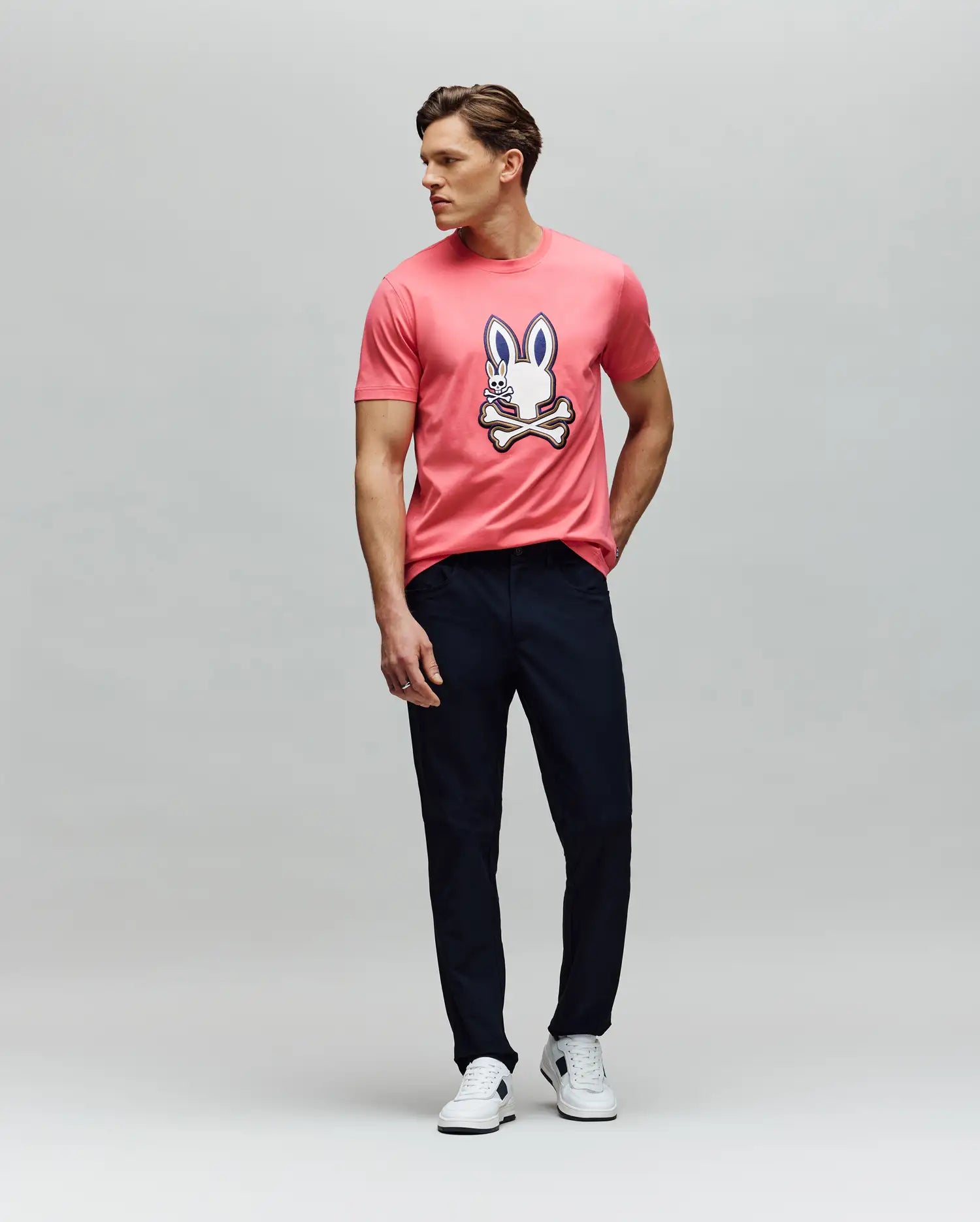 A man poses against a plain background in the Psycho Bunny MENS WADE GRAPHIC TEE (MT0200106), a pink 100% cotton shirt with an embroidered bunny and crossbones, styled with dark pants and white sneakers, one hand in his pocket. (5)