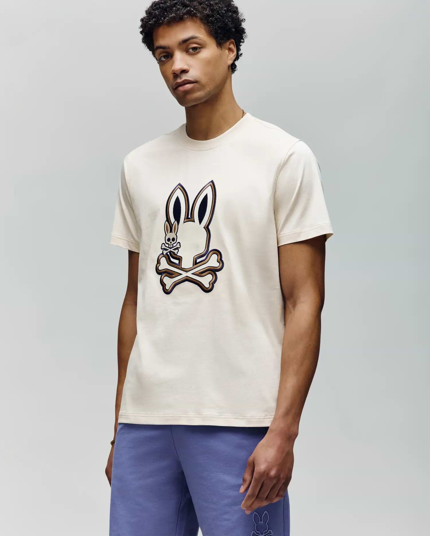 A man wearing the Psycho Bunny MENS WADE GRAPHIC TEE - MT0200106, a cream tee with a large embroidered bunny and crossbones on the front, paired with purple shorts, stands against a plain light gray background.
