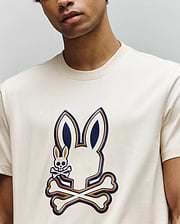 A man wears the Psycho Bunny MENS WADE GRAPHIC TEE - MT0200106, a cream 100% cotton shirt with a large stylized bunny head with crossbones and an embroidered bunny skull-and-crossbones on the front; his face is partially out of frame.