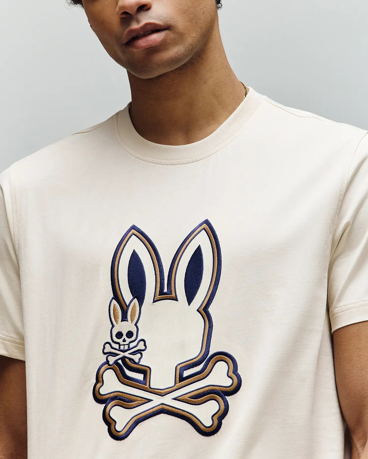 A man wears the Psycho Bunny MENS WADE GRAPHIC TEE - MT0200106, a cream 100% cotton shirt with a large stylized bunny head with crossbones and an embroidered bunny skull-and-crossbones on the front; his face is partially out of frame.