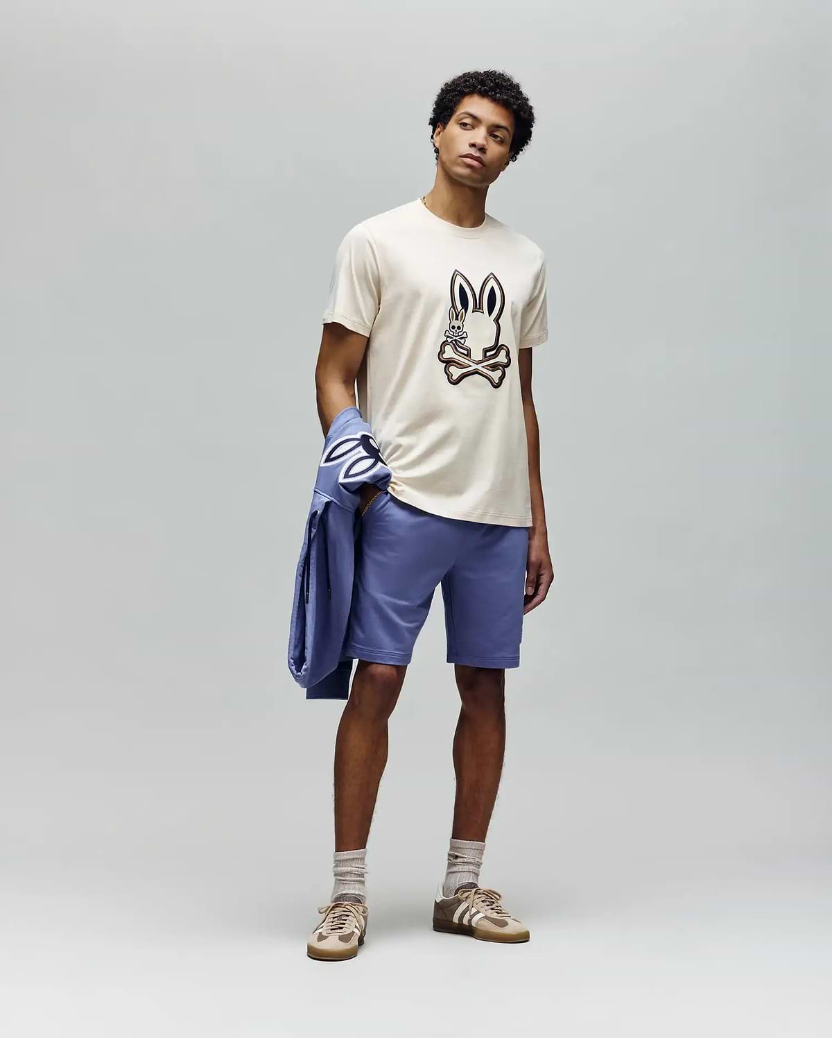 A man stands against a plain gray background wearing the Psycho Bunny MENS WADE GRAPHIC TEE - MT0200106 (beige, embroidered bunny face), lavender shorts, beige socks, tan sneakers, and holds a lavender jacket over his left arm.