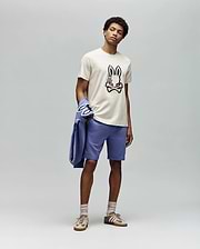 A man stands against a plain gray background wearing the Psycho Bunny MENS WADE GRAPHIC TEE - MT0200106 (beige, embroidered bunny face), lavender shorts, beige socks, tan sneakers, and holds a lavender jacket over his left arm.