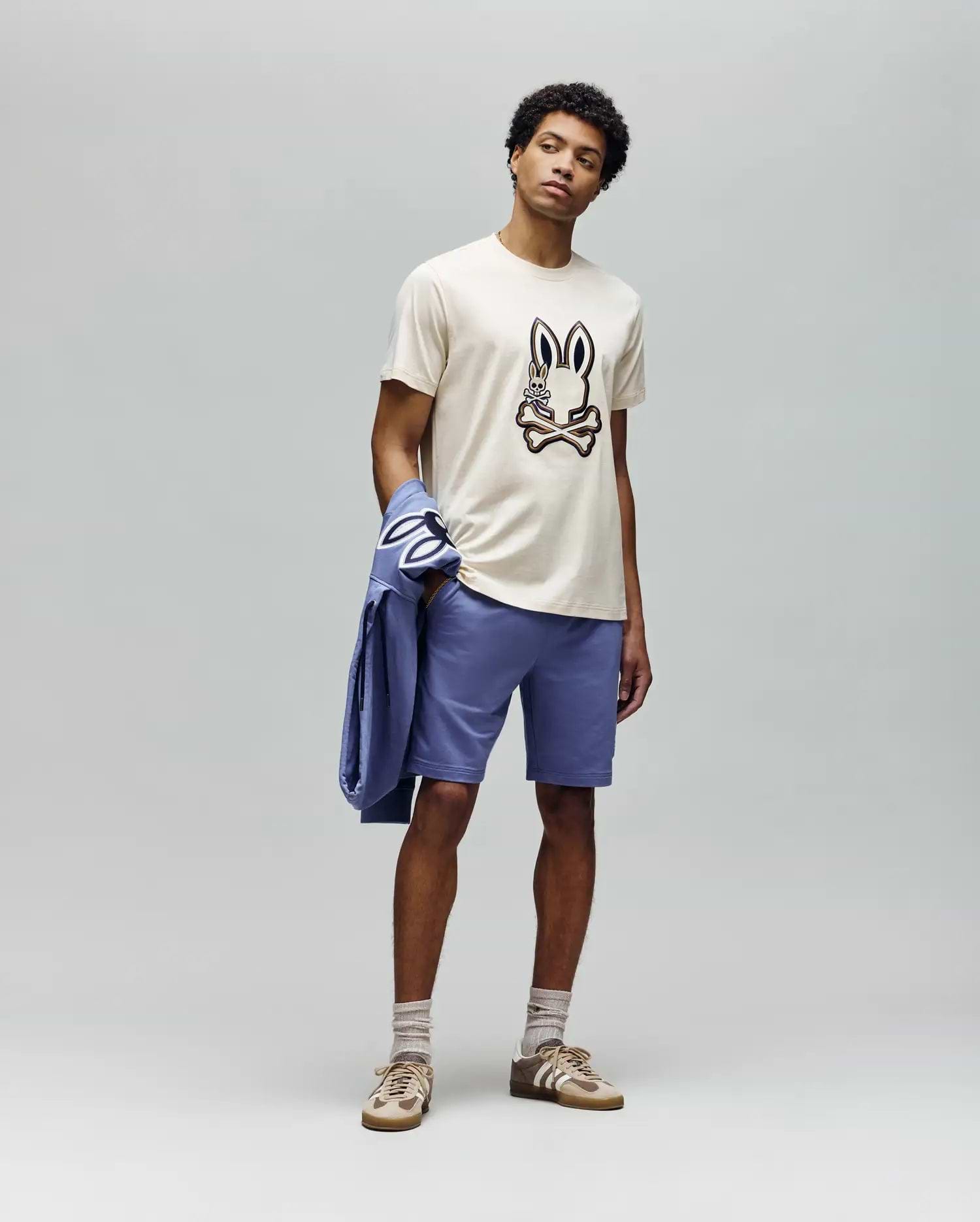A man stands against a plain gray background wearing the Psycho Bunny MENS WADE GRAPHIC TEE - MT0200106 (beige, embroidered bunny face), lavender shorts, beige socks, tan sneakers, and holds a lavender jacket over his left arm.