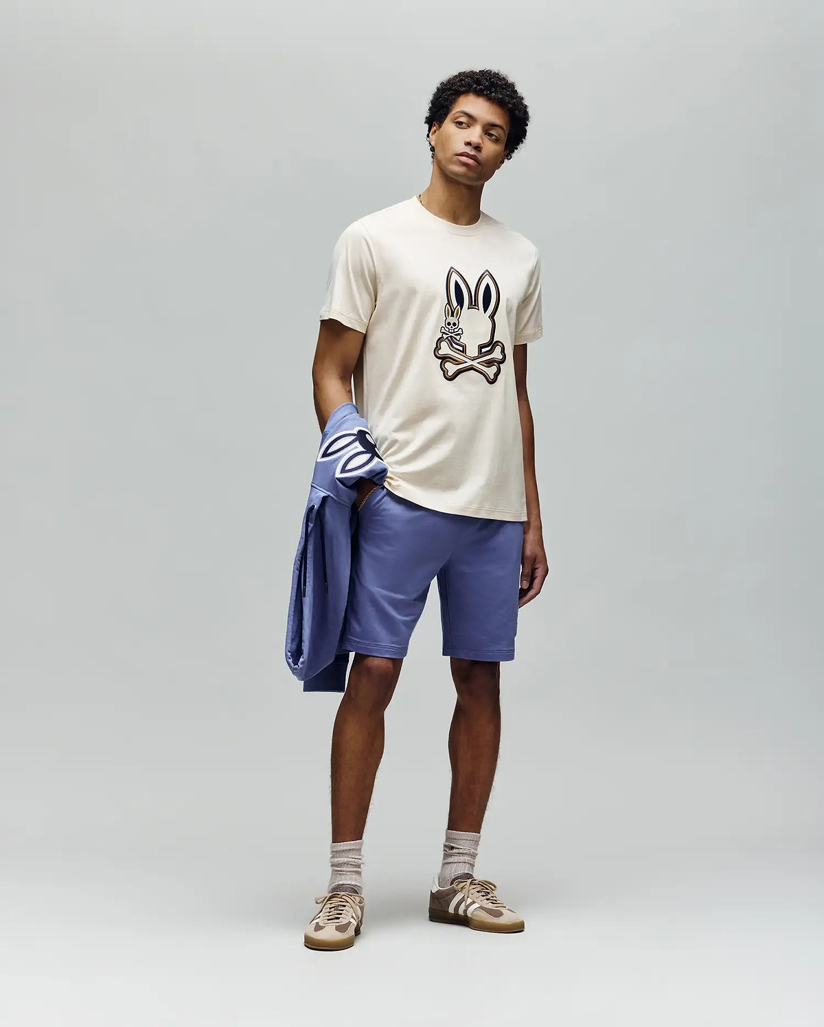 A man stands against a plain gray background wearing the Psycho Bunny MENS WADE GRAPHIC TEE - MT0200106 (beige, embroidered bunny face), lavender shorts, beige socks, tan sneakers, and holds a lavender jacket over his left arm.