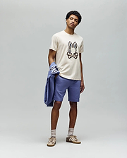 A man stands against a plain gray background wearing the Psycho Bunny MENS WADE GRAPHIC TEE - MT0200106 (beige, embroidered bunny face), lavender shorts, beige socks, tan sneakers, and holds a lavender jacket over his left arm.