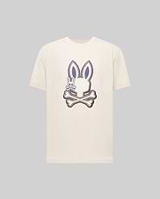 The Psycho Bunny MENS WADE GRAPHIC TEE (MT0200106) is a cream-colored shirt featuring a large bunny head over crossed bones with an embroidered bunny on the left, detailed in dark blue and beige on a plain background.