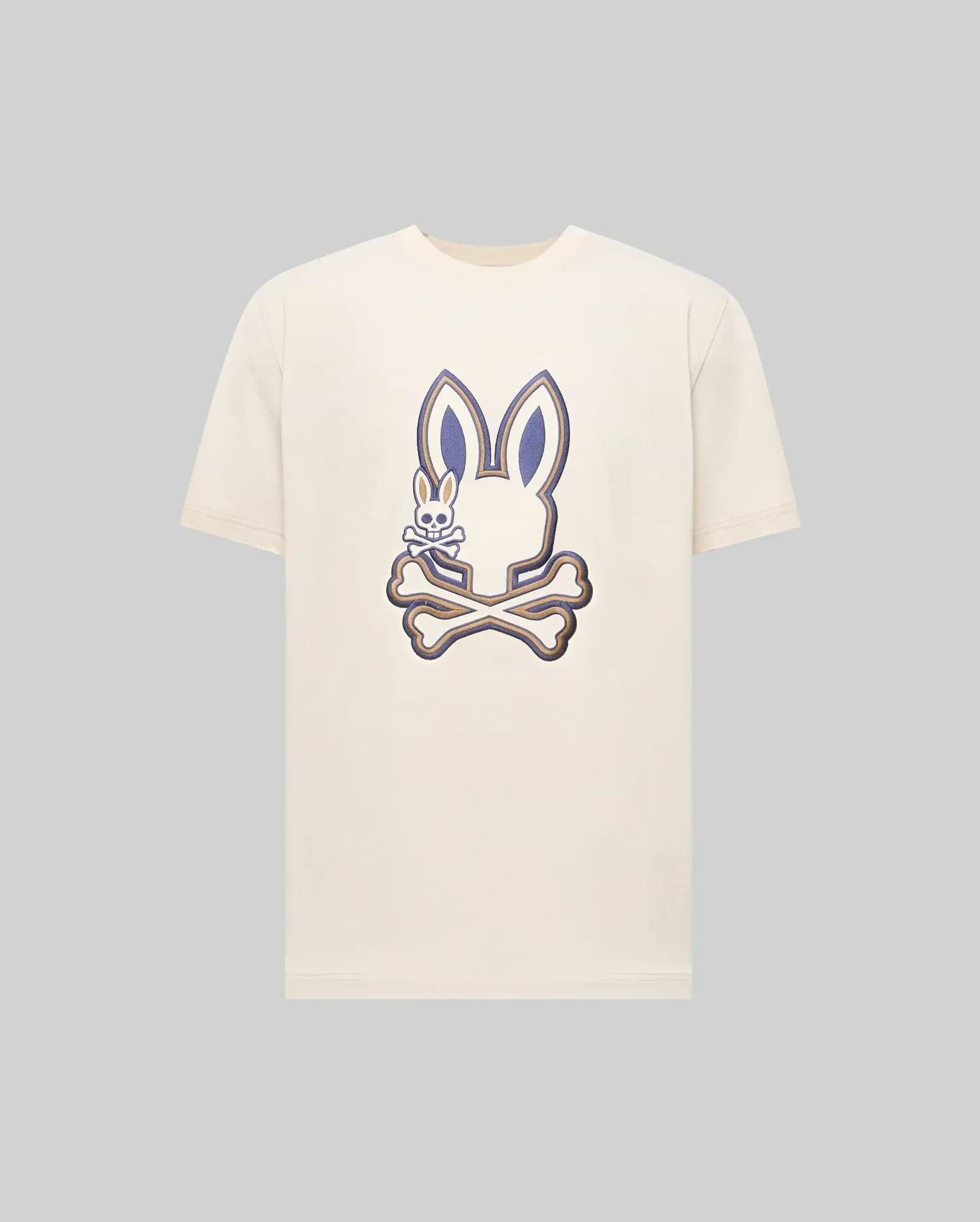 The Psycho Bunny MENS WADE GRAPHIC TEE (MT0200106) is a cream-colored shirt featuring a large bunny head over crossed bones with an embroidered bunny on the left, detailed in dark blue and beige on a plain background.