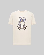 The Psycho Bunny MENS WADE GRAPHIC TEE (MT0200106) is a cream-colored shirt featuring a large bunny head over crossed bones with an embroidered bunny on the left, detailed in dark blue and beige on a plain background.