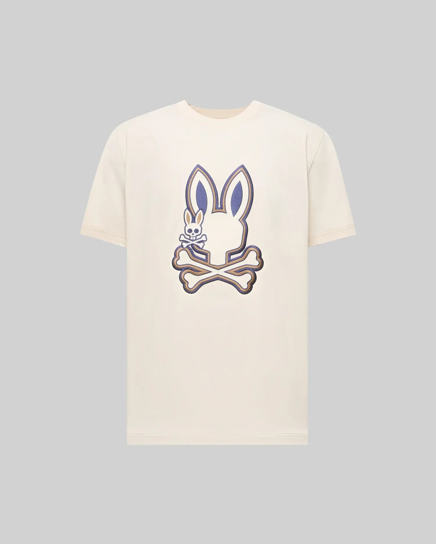 The Psycho Bunny MENS WADE GRAPHIC TEE (MT0200106) is a cream-colored shirt featuring a large bunny head over crossed bones with an embroidered bunny on the left, detailed in dark blue and beige on a plain background.