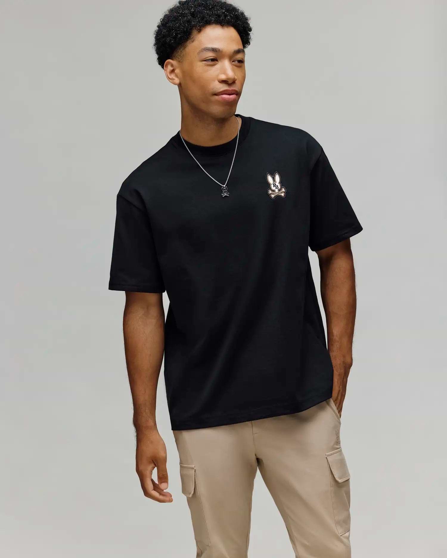 A young man stands against a light gray background, wearing the Psycho Bunny MENS WADE OVERSIZED FIT CHEST TEE (MT0200152), beige cargo pants, and a necklace, looking to the side with one hand in his pocket.