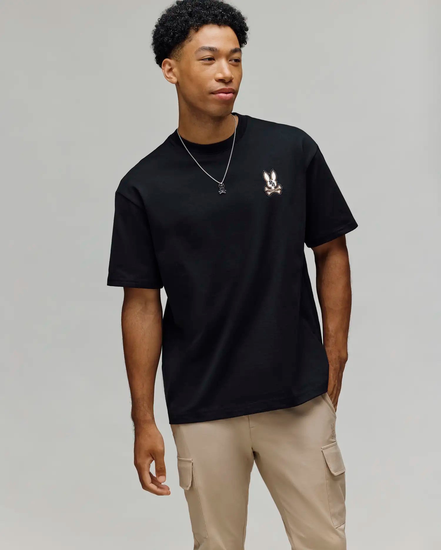 A young man stands against a light gray background, wearing the Psycho Bunny MENS WADE OVERSIZED FIT CHEST TEE (MT0200152), beige cargo pants, and a necklace, looking to the side with one hand in his pocket.