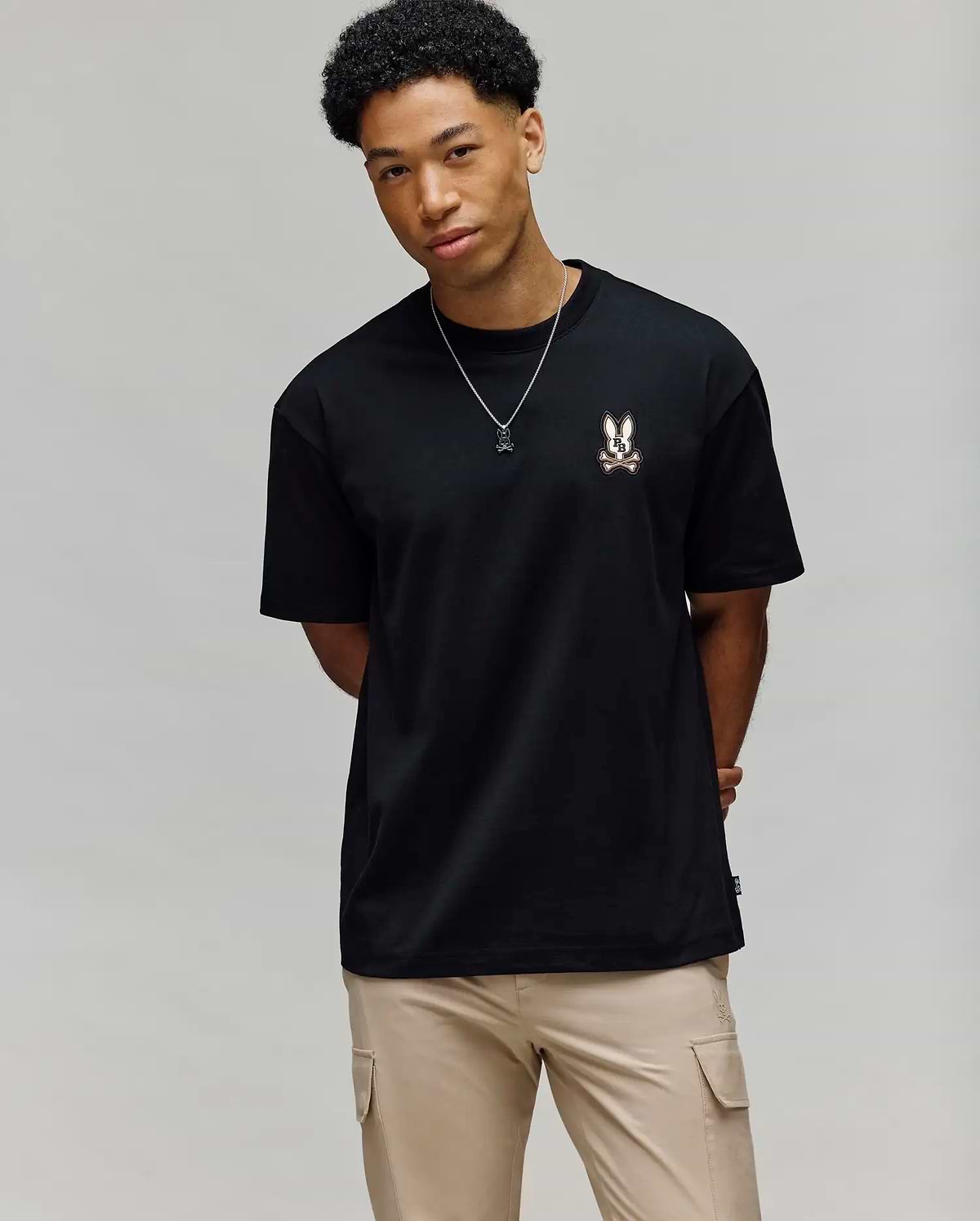 A young man with short curly hair wears the Psycho Bunny MENS WADE OVERSIZED FIT CHEST TEE (MT0200152) in black, featuring a small bunny graphic on the left chest, beige pants, and a necklace against a light grey background.