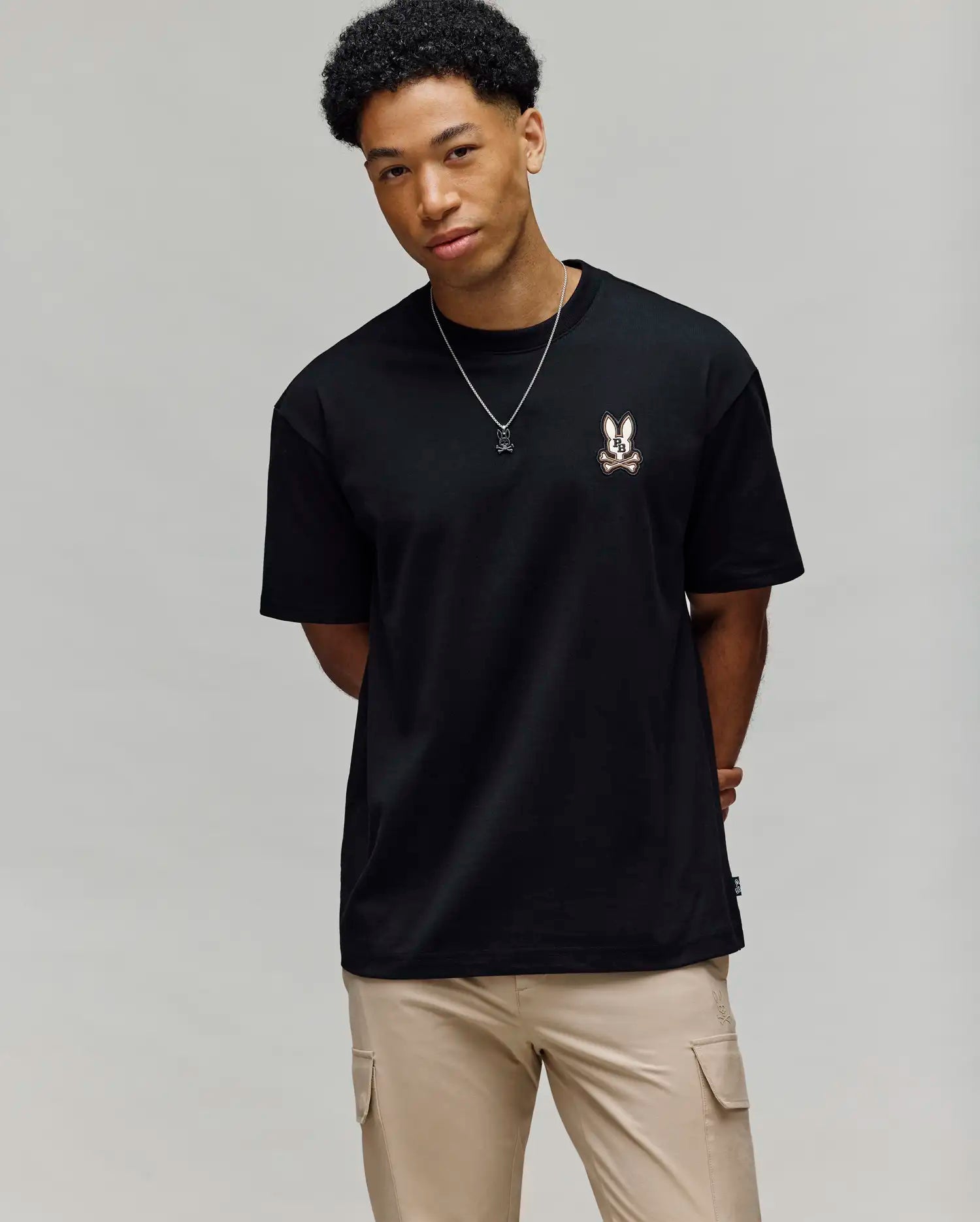 A young man with short curly hair wears the Psycho Bunny MENS WADE OVERSIZED FIT CHEST TEE (MT0200152) in black, featuring a small bunny graphic on the left chest, beige pants, and a necklace against a light grey background.